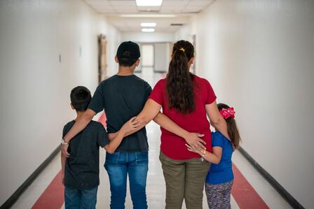 Father, mother, son, and daughter stand arm in arm, backs to the viewer, in a brightly lit hallway