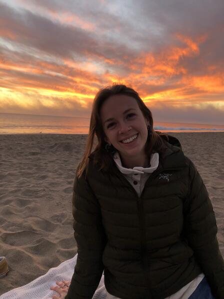 A woman in a coat sitting on a beach, with a colorful sunset in the background.
