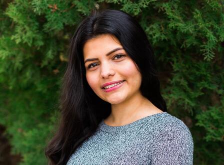 Kamala provided skills as the New Roots food access & education intern. Kamala smiles for the camera in front of evergreen shrubbery