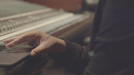 Closeup of Belly's hands working at a laptop perched on a mixing board in a recording studio