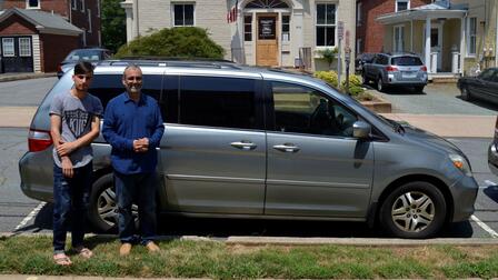 Two men stand in front of a silver minivan they have just received