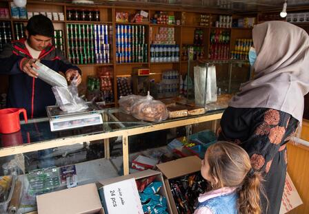 Noor and her daughter purchase food in a market in Afghanistan A man weights food while Noor and her daughter look on in an outdoor market in Afghanistan.