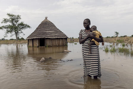 With water above her ankles, a mother holds her child and stands in front of her flooded home. 