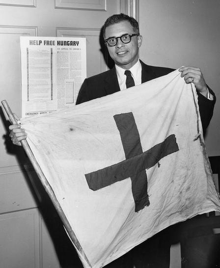 Leo Cherne in Hungary Leo Cherne poses for a picture holding a flag while standing in front of a sign that says "Help Free Hungary."