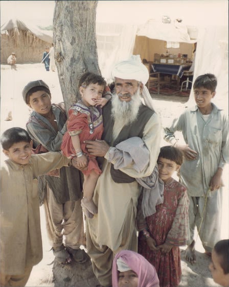An older man and a number of young boys, all Afghan refugees, pose for a photo outside, in front of a tree.