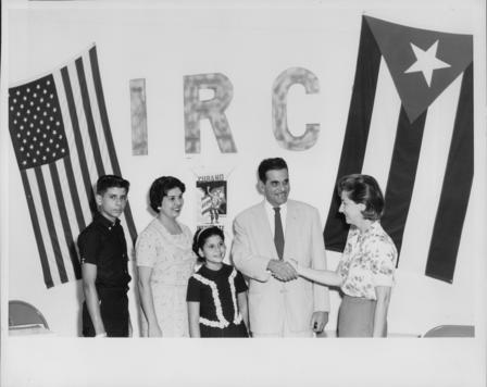 A Cuban family of four poses with an IRC staff member in front of a wall that says "IRC," next to a Cuban flag and a U.S. flag.
