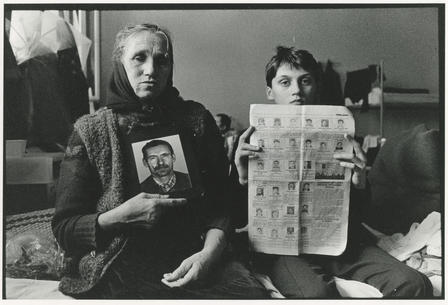 A woman and a young boy sit next to each other on a bed, holding up photos of the faces of men killed in conflict.