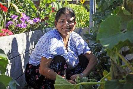 A woman crouches beside her garden plot, surrounded by greenery and flowers and smiling for the camera