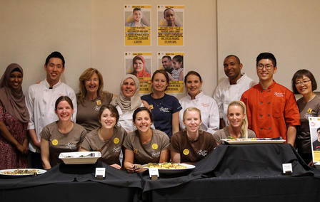 Spice Kitchen Incubator, a program of the International Rescue Committee, catered Journey to the Wasatch. Volunteers, staff and entrepreneurs pictured here. Staff members and chefs of the Spice Kitchen Incubator sit and stand behind a table with smiles.
