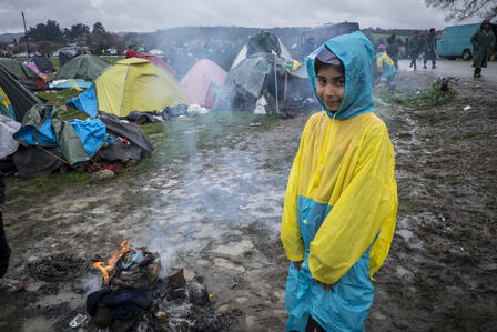 A Syrian girl in a raincoat stands by a campfire in a refugee settlement in Greece