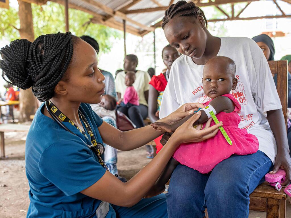 Eine Ärztin im blauen Kittel untersucht ein Kind auf Unterernährung während es auf dem Schoß der Mutter sitzt