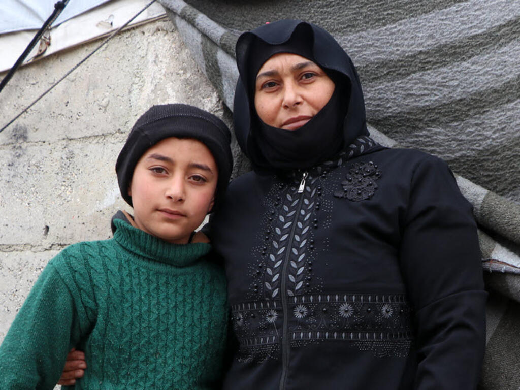 A Syrian woman stands with her son in a displacement camp. They look directly at the camera, bundled in dark, thick winter clothing. 