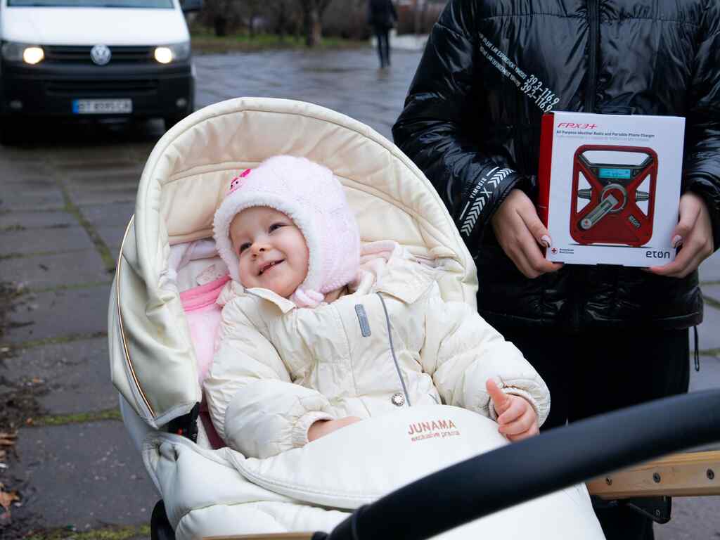 A young girl wrapped in warm winter clothes smiles in her stroller. Her caregiver is holding a winter kit from the IRC.
