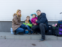 Parents, two children and a cat pose for a photo. Besides them are suitcases packed with their belongings.