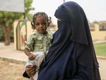 A woman in a burqa carrying her daughter who is looking at the camera in a green shirt