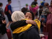 A woman smiles with her eyes closed as she embraces another person inside a busy arrivals hall, surrounded by others reuniting with loved ones.