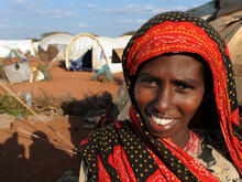 A smiling young woman stands outside a tent in Dadaab refugee camp in Kenya on a sunny day,