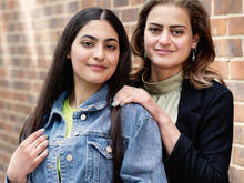 A mother stands behind her daughter with her hand on her shoulder. Both look at the camera. 