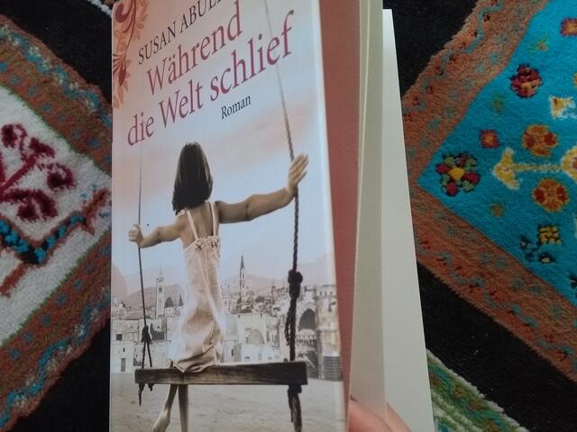 Ein Buch liegt auf einem bunten Teppich und wird von einer Hand offen gehalten