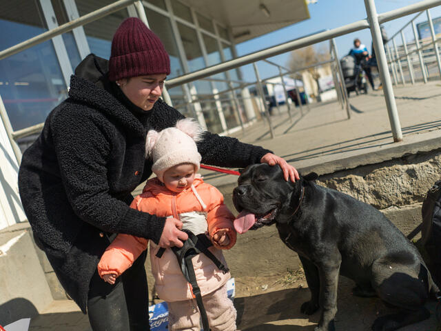 A mom stands with her young daughter and a dog beside them