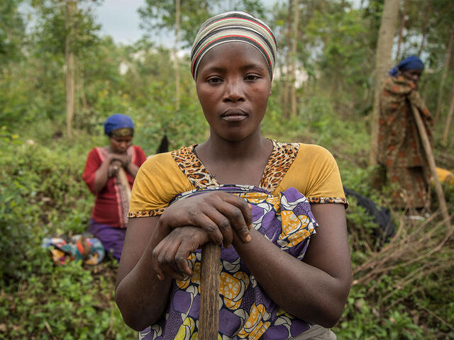 A woman pauses her agricultural work to pose for a photo in North Kivu, DRC. Two other women continue to work behind her.