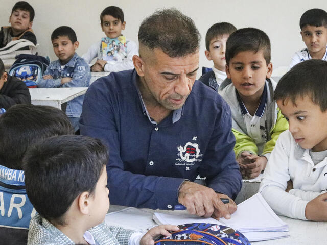 A man sits at a table surrounded by children, who watch him instruct with a notebook and pen.