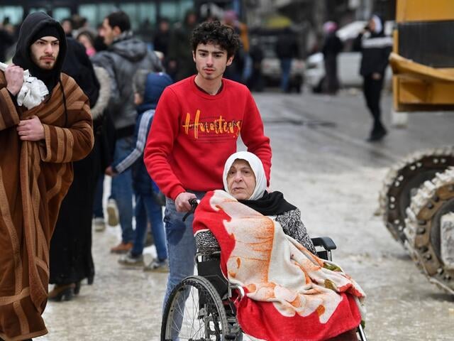 Two young Syrian men pushing an older woman in a wheelchair out of the disaster area.