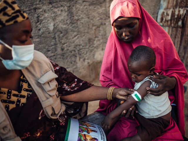 An IRC nutrition officer teaches a mother how to screen her child for signs of malnutrition using MUAC tape in Nigeria. The malnutrition wraps the tape around a boys arm while the mother looks on.