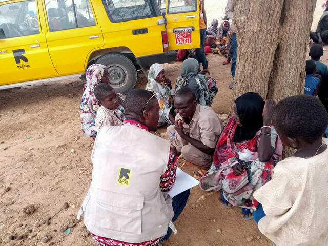 An IRC staff speaks to a group of Sudanese refugees who have recently arrived in Chad to assess their needs. The conversation takes place outside, with a number of other refugees in the background.