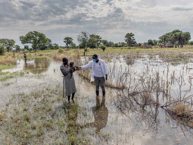 Abuk Deng holds her 4 year-old daughter in her arms as they walk away from their flooded home while an IRC nutrition officer walks alongside them.