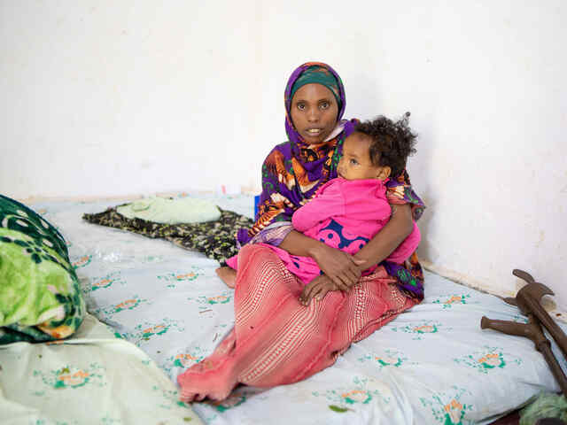 Eine Frau sitzt auf einer Matratze und hält ein Kleinkind im Arm, neben ihr sind Krücken.