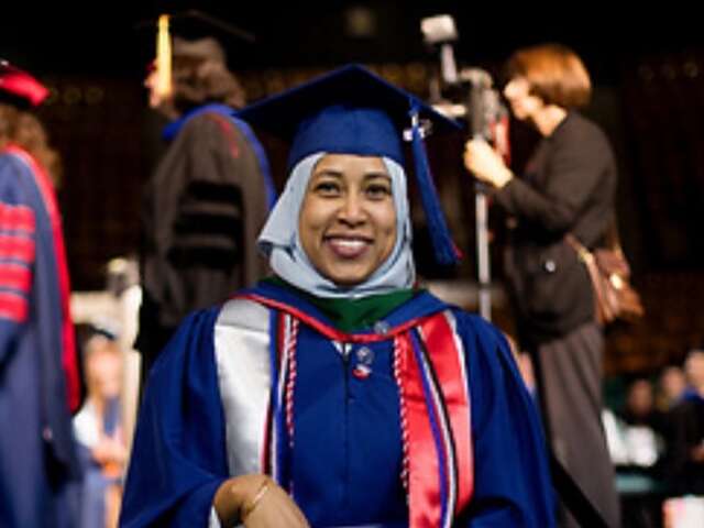 Selma Hamed is pictured smiling at graduation with blue cap, hijab, and gown with red honor cords 