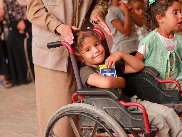 A young Palestinian girl in a wheelchair holds a pack of colored pencils in her arms and smiles.