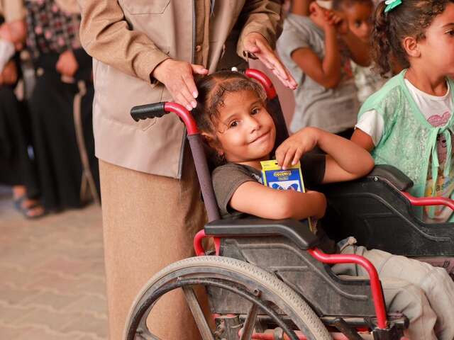 A young Palestinian girl in a wheelchair holds a pack of colored pencils in her arms and smiles.