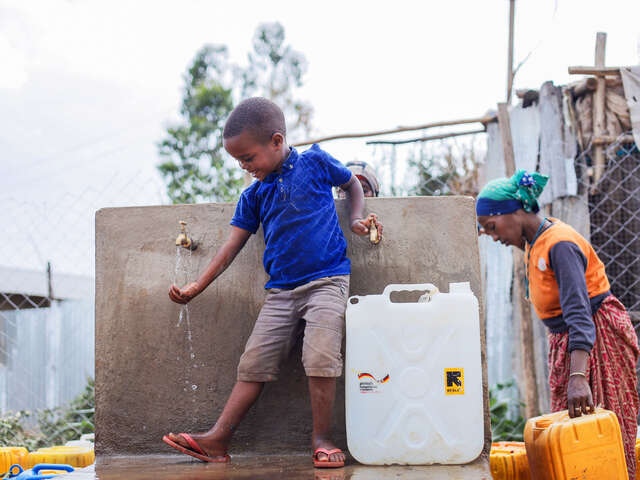 En pojke känner på vattnet vid en vattenstation i ett flyktingläger i Kenya, som RESCUE installerat. I bakgrunden syns hans mamma som bär två stora vattendunkar.