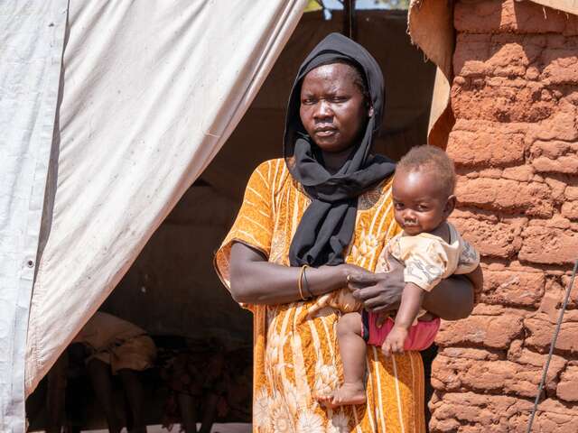 En mamma håller sin lilla dotter på höften och poserar för ett foto utanför sitt hem i Sydsudan.