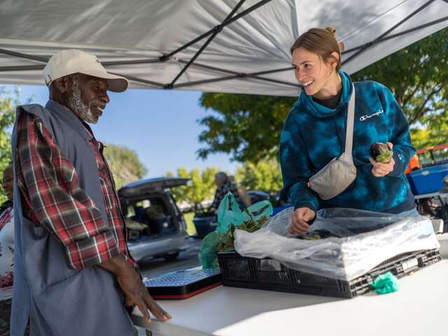 Omar, left, a New Roots farmer stands at his farmers market stall across from a customer holding an eggplant he grew locally.