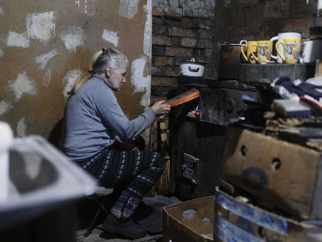 En kvinna lägger in ved i en vedspis i sitt halvt förstörda hem. 