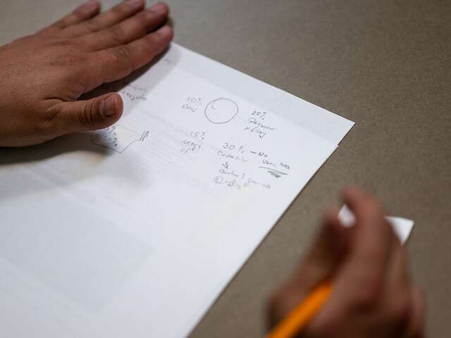 Close up shot shows an IRC VESL participant's hands as they take notes on a sheet of paper, pencil held in their right hand. Notes reflect the financial literacy lesson.