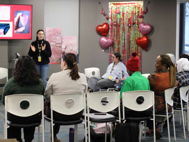 IRC clients sit facing with their backs to the camer as a team member presents information on menstruation, there are Valentine's decorations hanging on the wall of the room. 