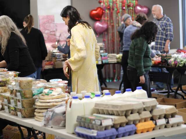 IRC volunteers and staff in the background work to organize tables filled with various grocery items; the foreground shows cartons of eggs, packaged sandwiches, and other food items.