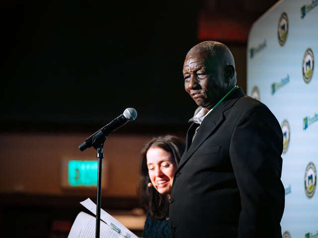 Albert, dressed in a suit coat, stands at a profile view, centered, in front of a microphone speaking to a Sundance audience in 2026.
