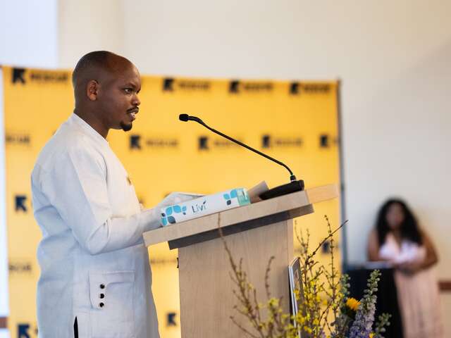 Profile of Emile Ndahunga from his right side, dressed in a white shirt, standing at a podium with a microphone. Background includes a banner with the International Rescue Committee logo.