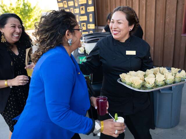 Wendy Juarez, owner of Prime Corn and the caterer for IRC's Rebuilding Lives Elevating Futures dinner, stands to the right holding a tray of guacamole with chips greeting two female guests, one in blue and another in black.