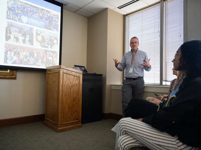 A Zions Bank recruiter stands while presenting to refugee youth participants of the IRC's Future Ready Summer Program, a slide deck is projected on the screen to the left of the presenter.