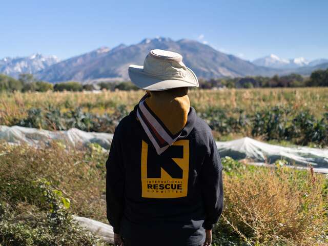 IRC team member stands in a New Roots field facing the Wasatch Mountains wearing a black sweater with the International Rescue Committee logo on the back and a wide brim beige hat.