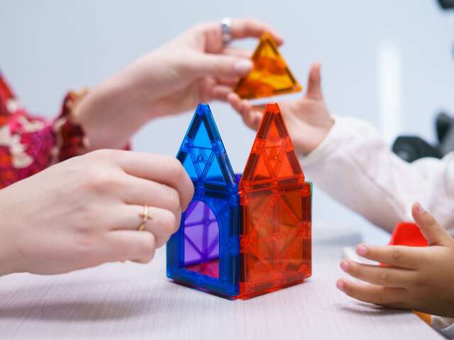 An IRC team member offers a moment of play to a child. Hands are present building connector blocks into the form of a house on a tabletop. 