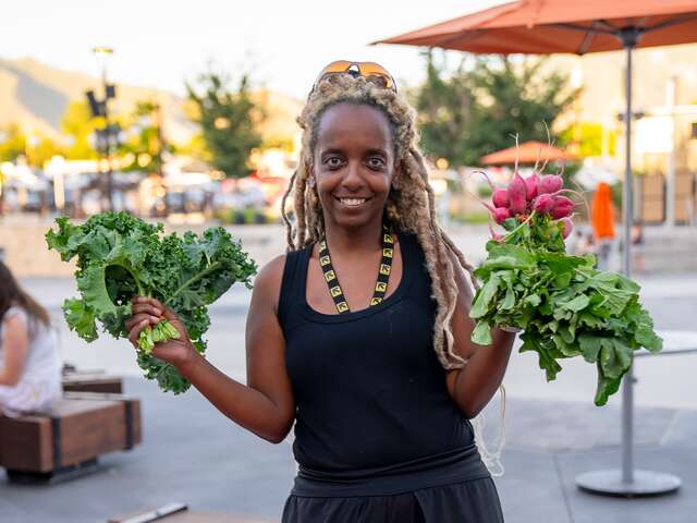 Selam, dressed in black, hair up wearing an IRC lanyard, holds bunches of fresh produce at her Rollin' Harvest booth at the Millcreek Farmers Market. 
