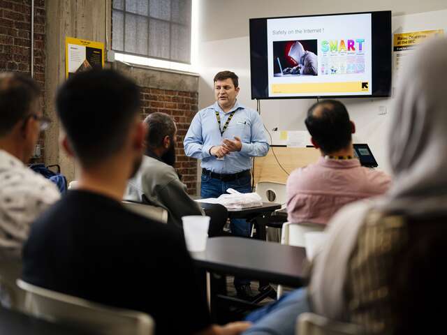 IRC teacher stands in the center background facing the class (and camera) with a slide about internet safety behind him. Class participants face him (away from the camera) learning about digital inclusion topics.