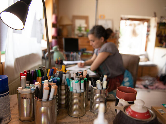 Blurry image with a close up of paint brushes and pencils with an outline of the artist Diala drawing at her desk.